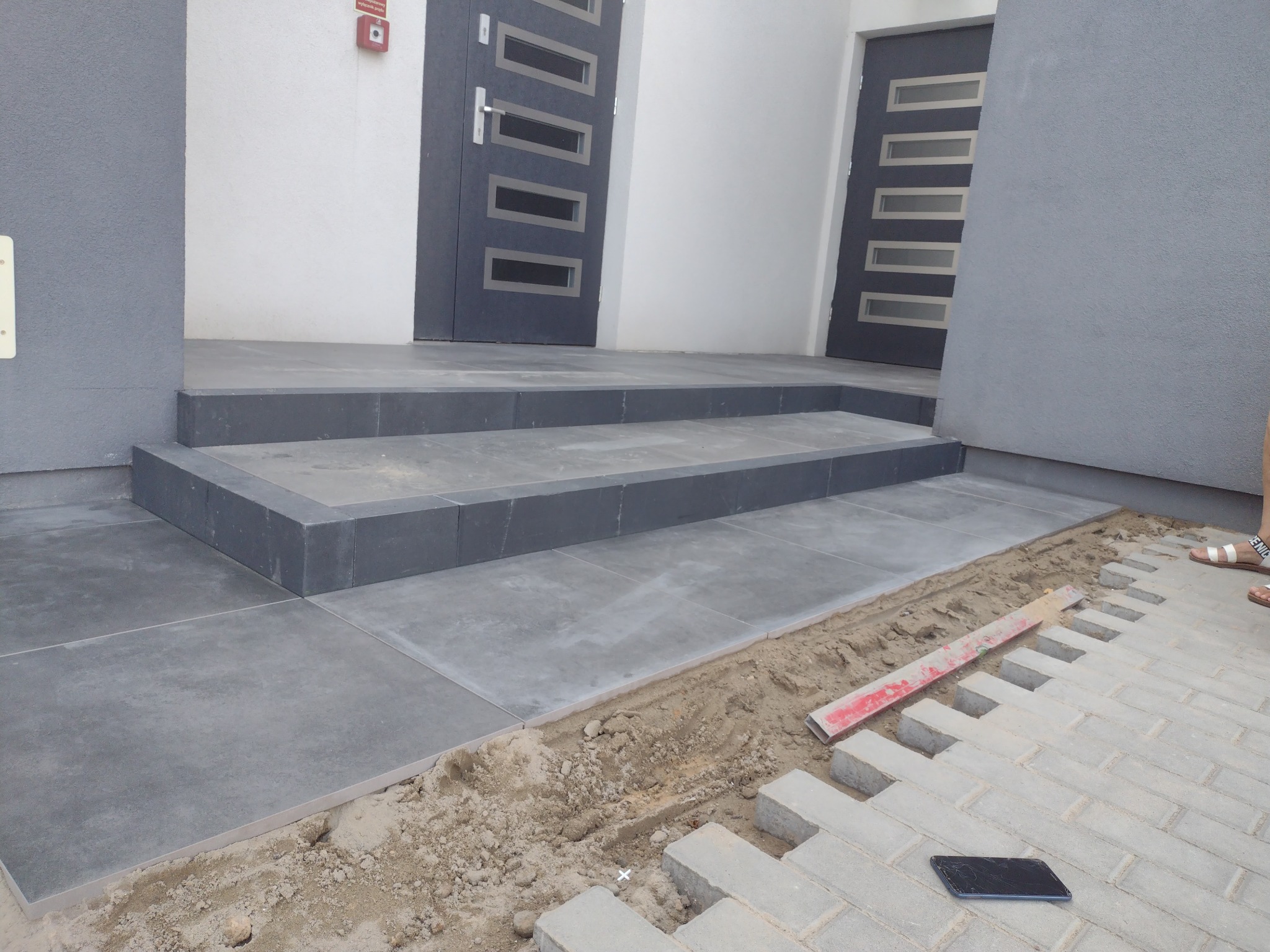 Schody wejściowe wyłożone szarymi płytami prowadzące do dwóch nowoczesnych drzwi; w tle fragment układanej kostki brukowej z poziomicą i telefonem.