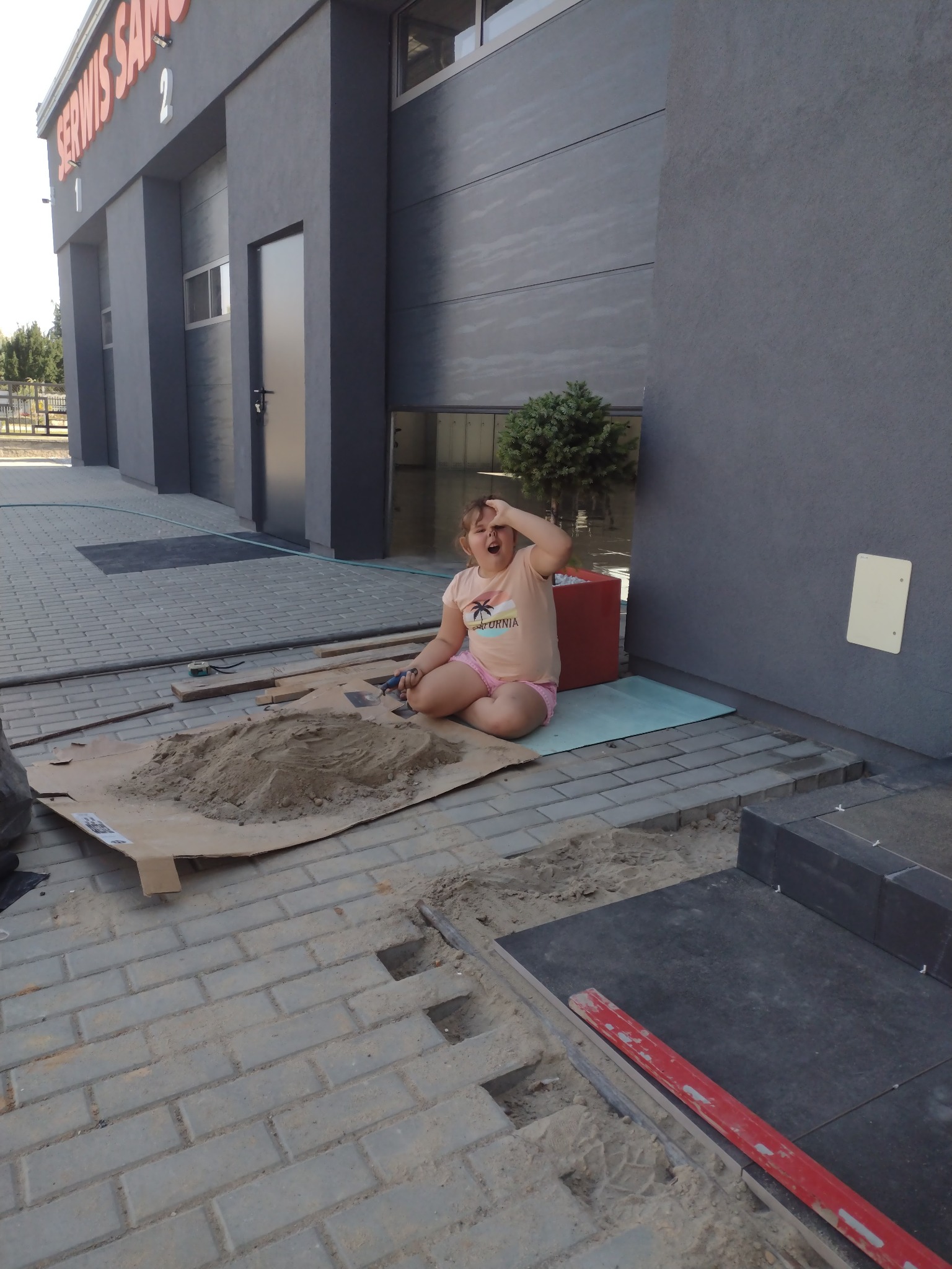Dziewczynka siedzi na kartonie obok kopca piasku podczas układania kostki brukowej przed budynkiem z szarą fasadą i napisem 'SERWIS SAM'.