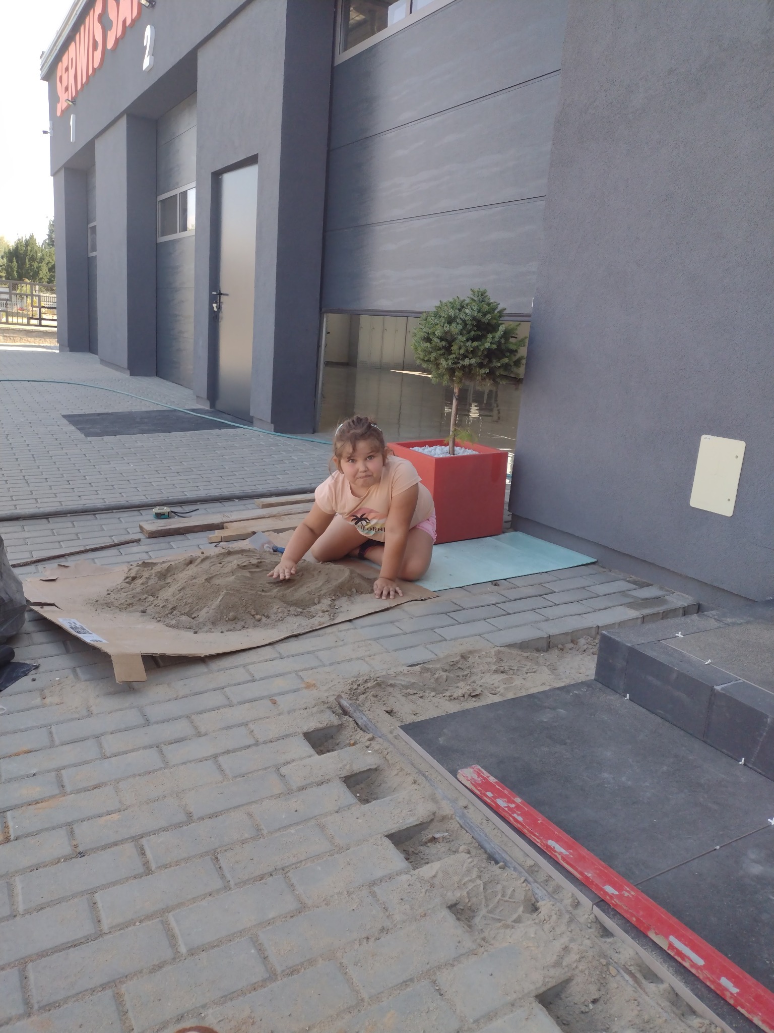 Dziewczynka bawi się piaskiem na tekturze podczas prac brukarskich, widoczne nieułożone kostki, poziomica i budynek w tle.