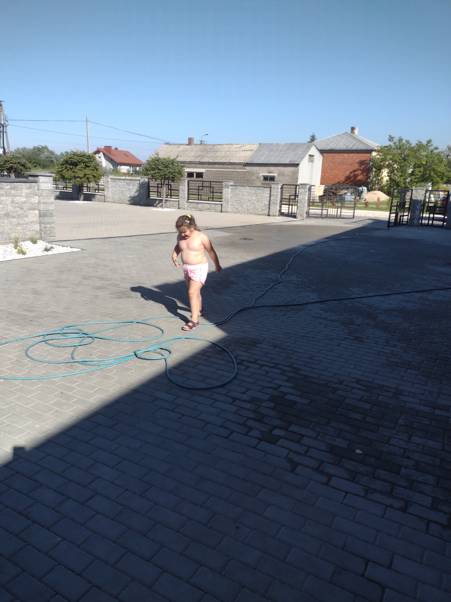 Dziewczynka idzie po nowo ułożonej, szarej kostce brukowej, na której leży zwinięty niebieski wąż ogrodowy. W tle widoczne ogrodzenie z szarej kostki i brama.