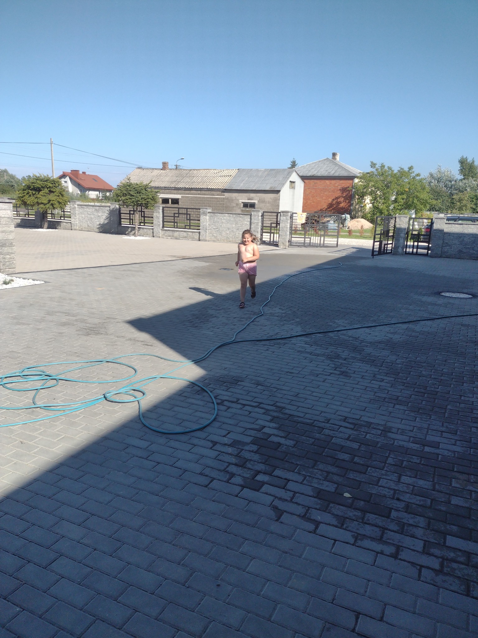 Dziewczynka biegnie po nowo ułożonej, szarej kostce brukowej na podjeździe, widoczny błękitny wąż ogrodowy i ogrodzenie z kamiennymi słupkami i metalowymi przęsłami.