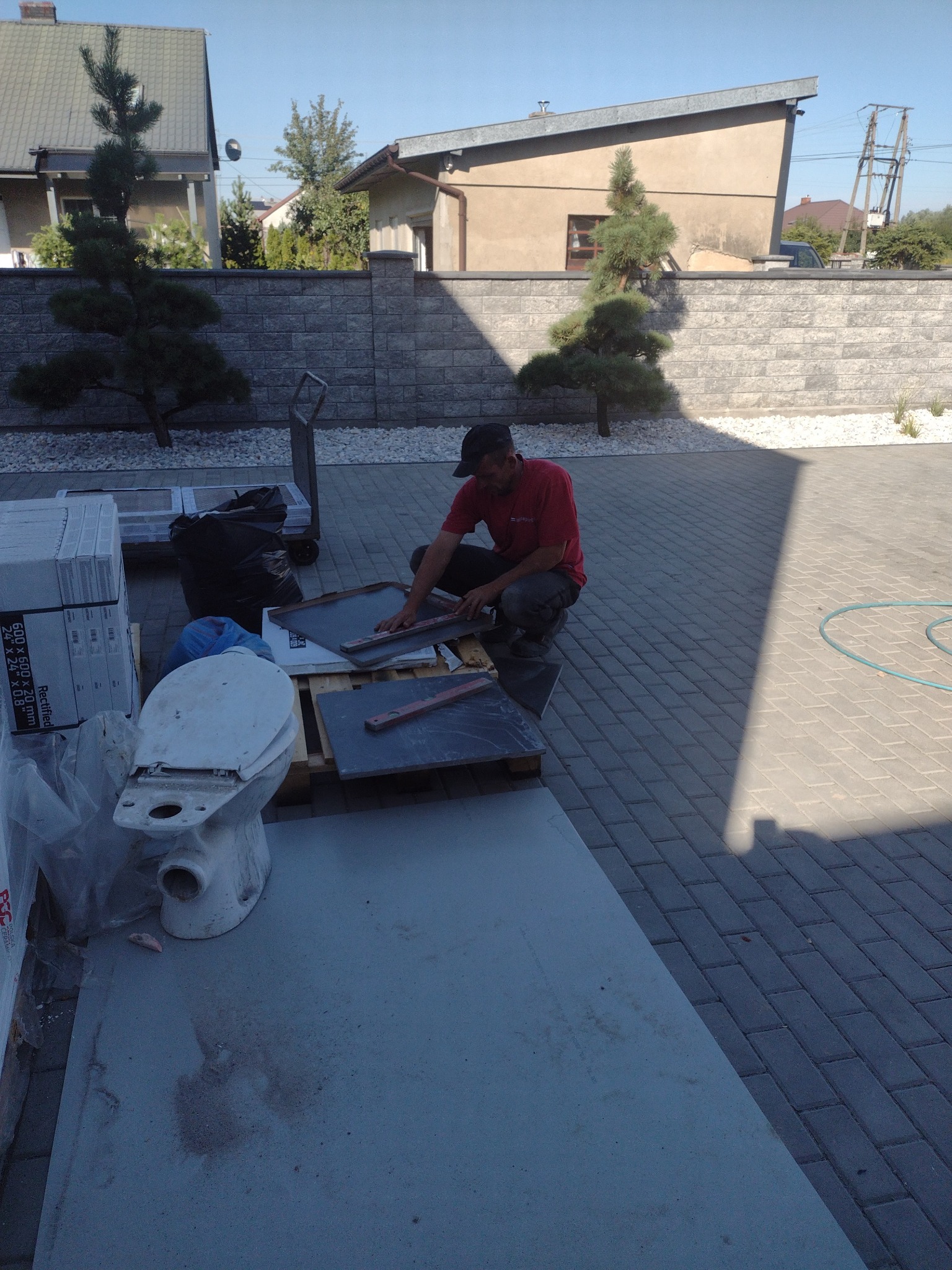 Pracownik w czerwonej koszulce docina ciemne płyty kamienne na palecie obok kostki brukowej, z toaletą w tle, na zewnątrz budynku.