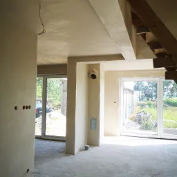 Surowe wnętrze po nałożeniu jasnych tynków, widoczne niedokończone schody drewniane, puszki elektryczne i okna przesuwne z widokiem na zieleń.