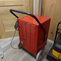 Czerwony, przemysłowy osuszacz powietrza na kółkach, stojący na panelach podłogowych, obok widoczny odkurzacz przemysłowy i drzwi w tle.