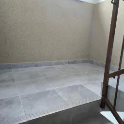 Wnętrze klatki schodowej z szarymi płytkami na podłodze i ścianami pokrytymi beżowym tynkiem strukturalnym. Widoczna metalowa balustrada schodowa w kolorze brązowym.