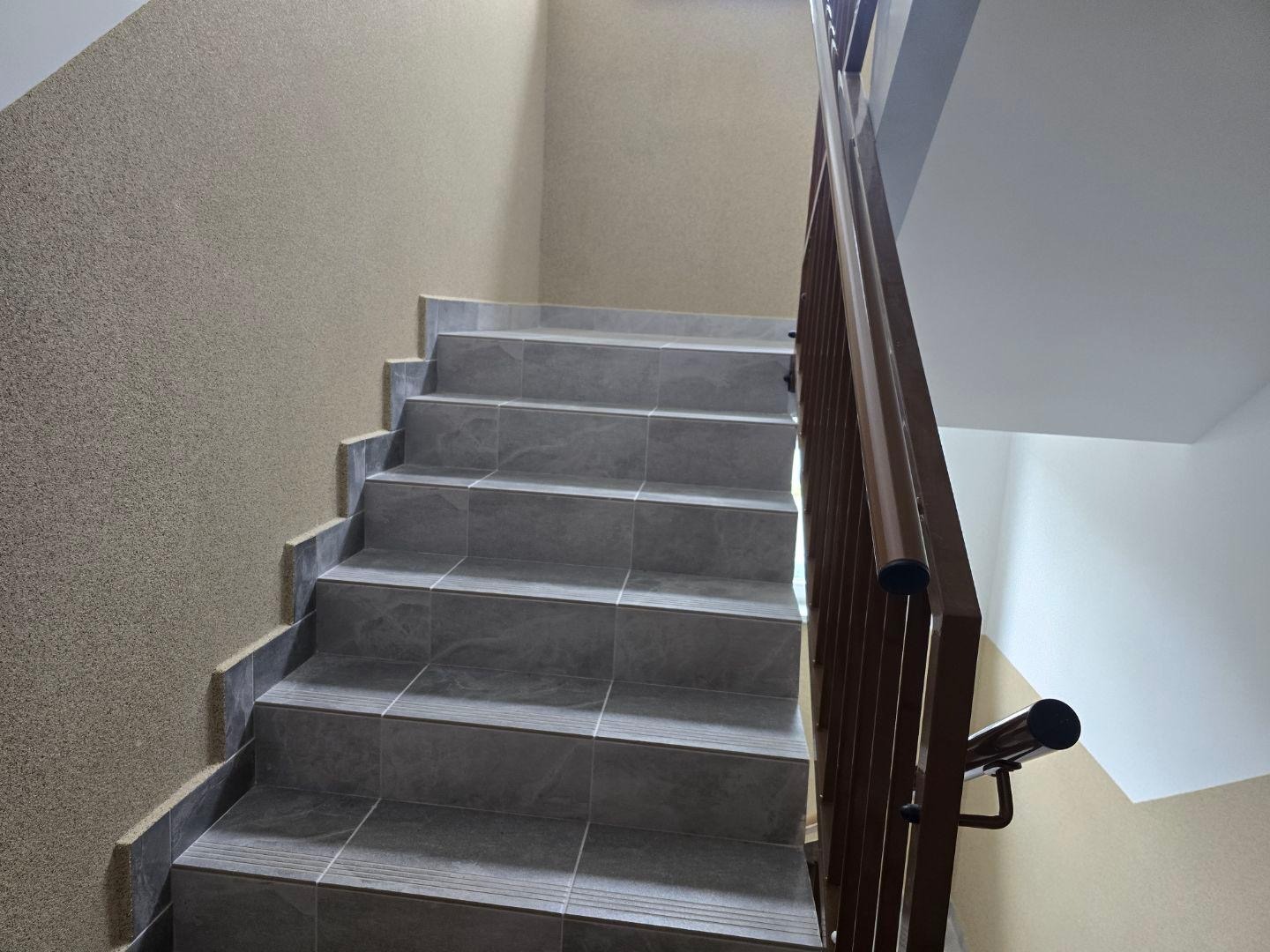 Wewnętrzne schody z szarych płytek z brązową balustradą, widok z dołu. Ściany w odcieniach beżu, faktura ziarnista. Ujęcie podkreśla detale wykończenia.