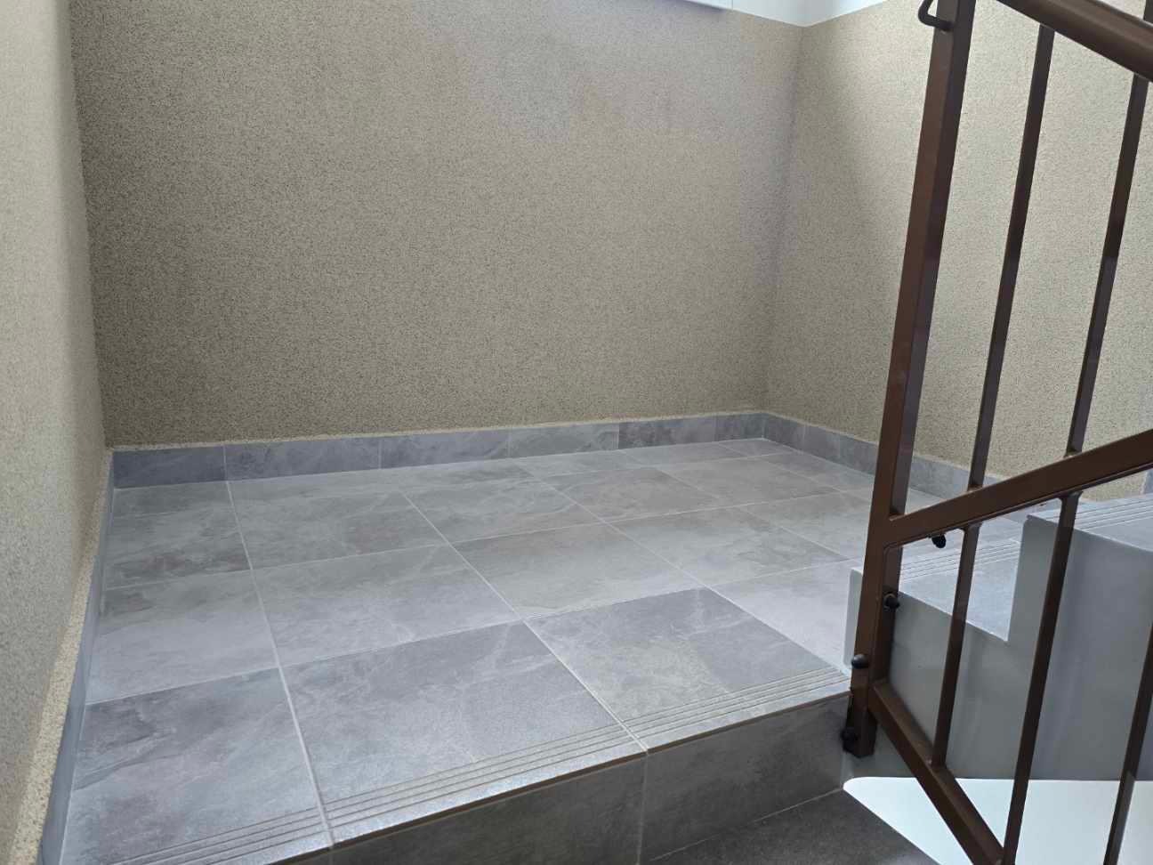 Wnętrze klatki schodowej z szarymi płytkami na podłodze i ścianami pokrytymi beżowym tynkiem strukturalnym. Widoczna metalowa balustrada schodowa w kolorze brązowym.