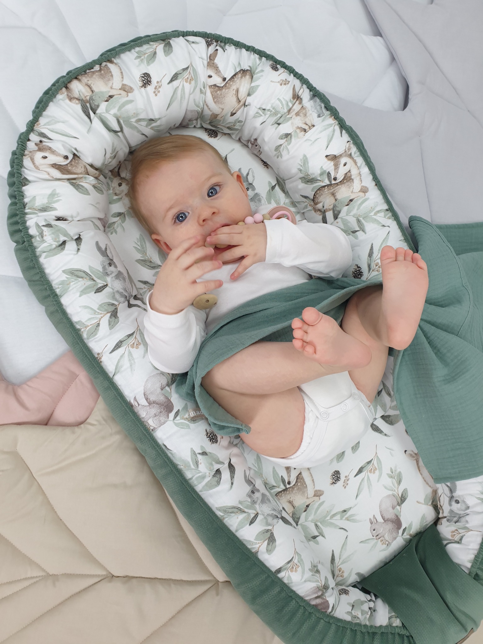 Niemowlę leżące w kokonie z motywem leśnych zwierząt, otulone zielonym kocykiem. Widoczny fragment kołderki w beżowym kolorze.
