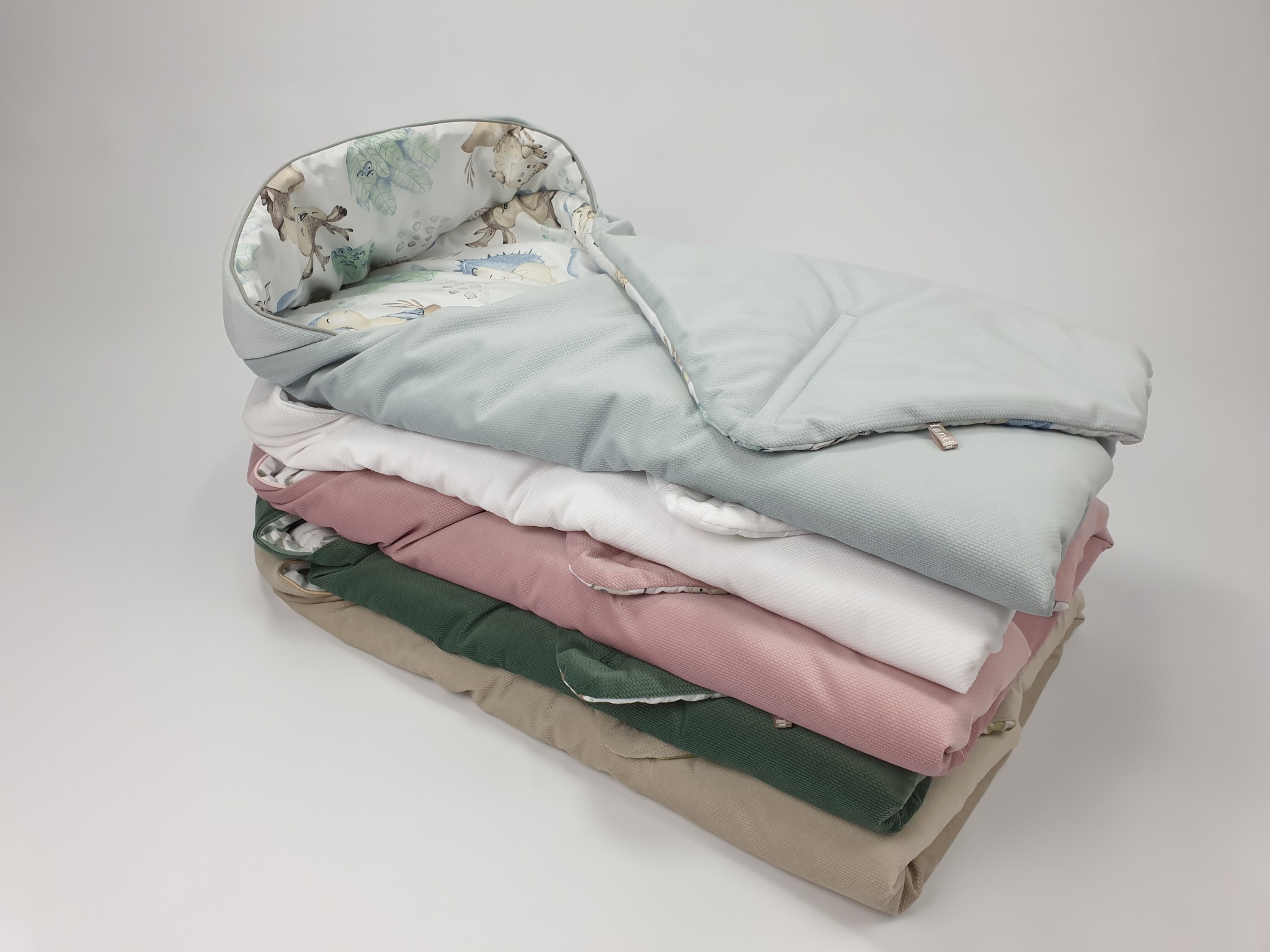 Stos pokrowców na śpiworek dziecięcy w pastelowych kolorach: beż, zieleń, róż, biel i błękit, z wzorem zwierząt w środku, na jasnym tle.