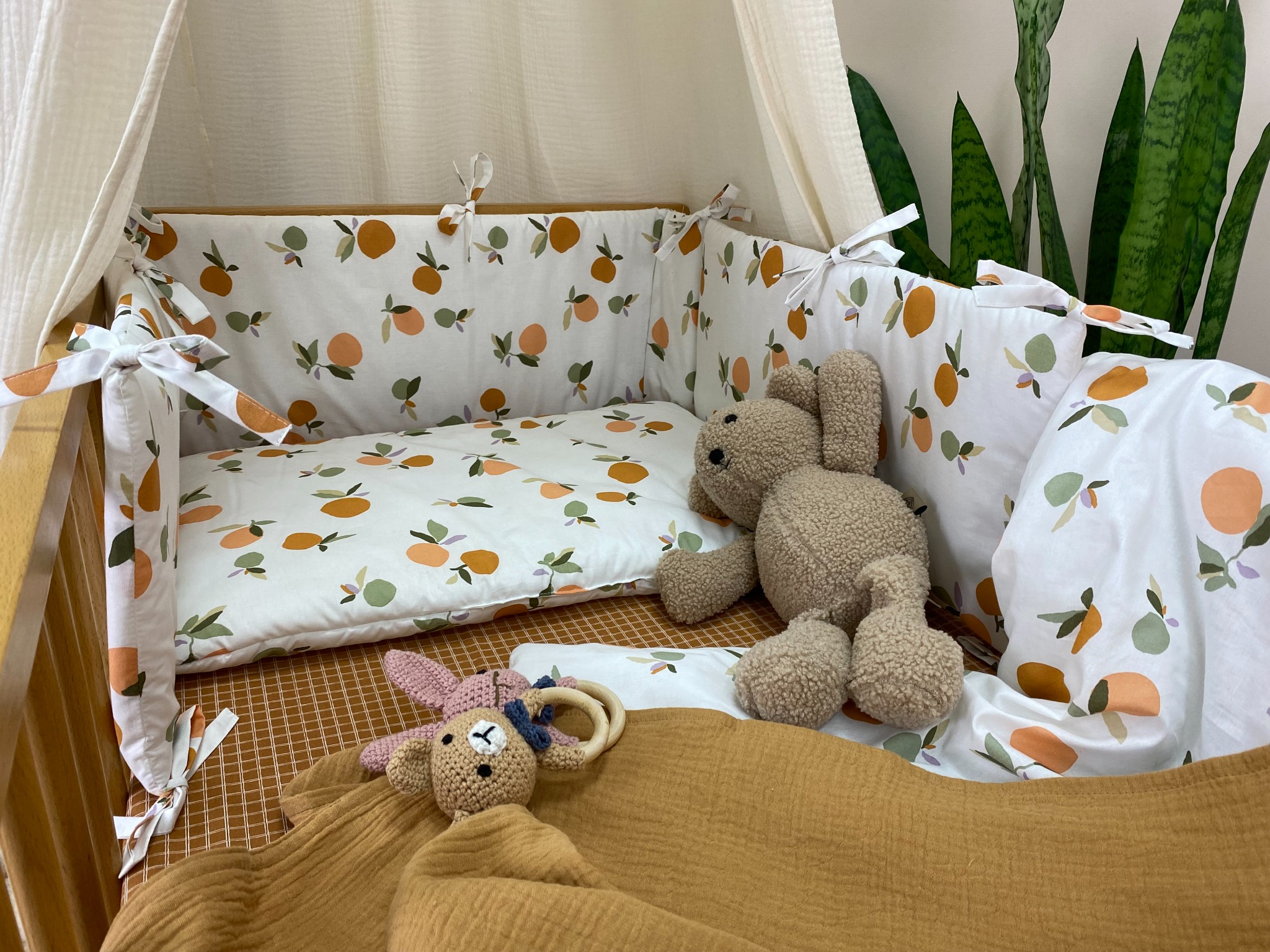 Łóżeczko dziecięce z kompletem pościeli w pomarańczowe wzory, otulaczem i zabawkami, widok z góry. W tle zielona roślina doniczkowa.
