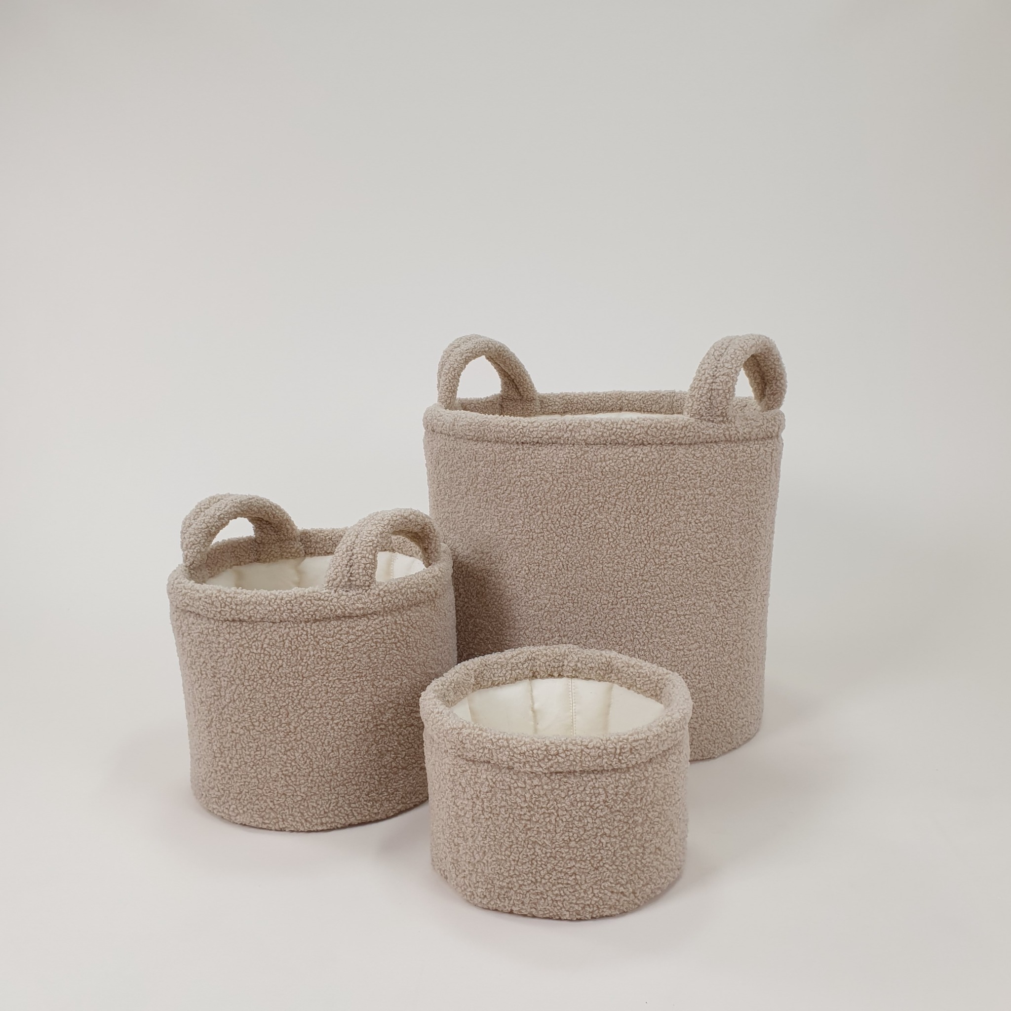 Trzy beżowe, pluszowe kosze z uchwytami, różnej wielkości, na białym tle. Widoczne przeszycia i kremowe wnętrze.