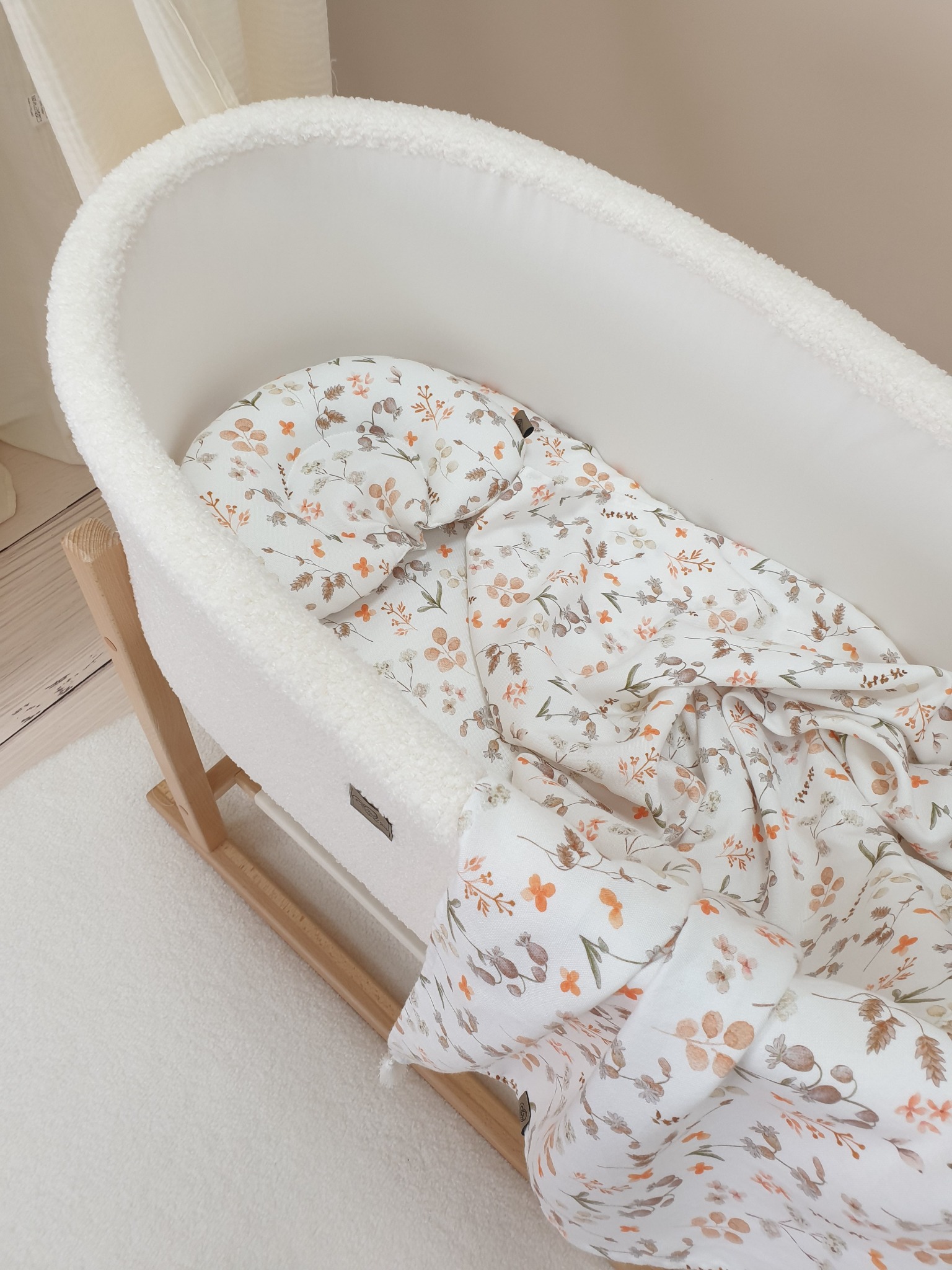 Biała kołyska z miękkim wykończeniem i pościelą w kwiatowy wzór, leżąca na puszystym dywanie.