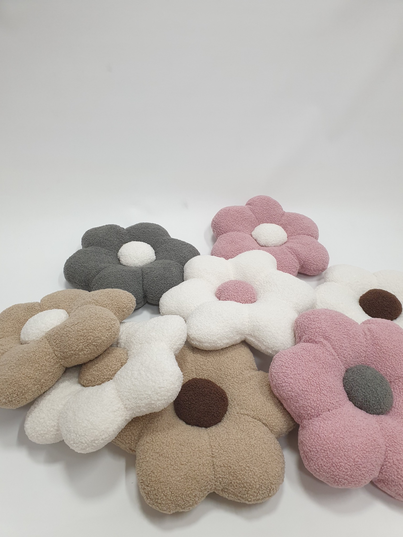 Różnokolorowe, pluszowe poduszki w kształcie kwiatów z okrągłymi środkami, leżące na białym tle. Widać odcienie szarości, różu, beżu i bieli.