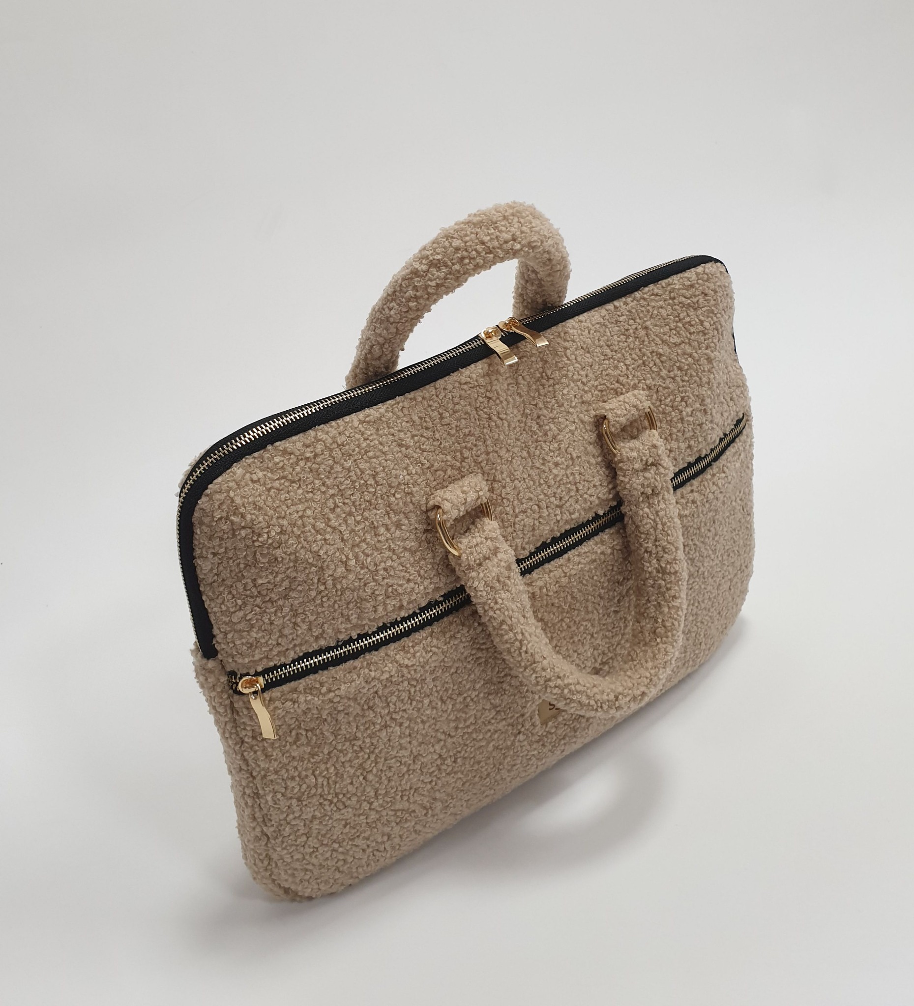 Beżowa torba z materiału typu baranek, z czarnymi zamkami i złotymi detalami, na białym tle. Widoczne uchwyty i zamek błyskawiczny.