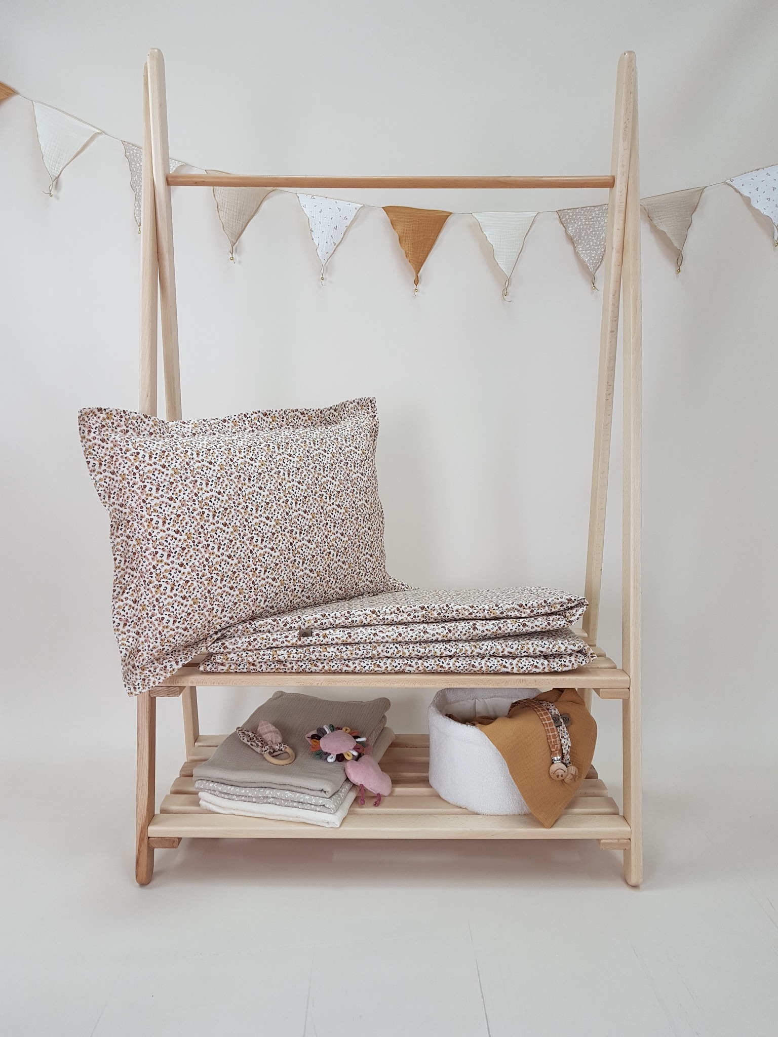 Jasna pościel w drobne kwiatki, ułożona na drewnianym stojaku z półkami. W tle girlanda z trójkątnych proporczyków. Przytulna aranżacja dla dziecka.