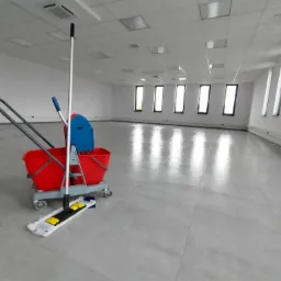 Wózek z mopem i wiadrem na tle świeżo umytej, błyszczącej podłogi w pustym biurze z rzędem okien.