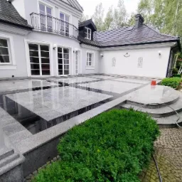 Elegancki taras z szarej kostki brukowej ułożonej we wzór szachownicy, przylegający do białego, dwupiętrowego domu z balkonem i zadbanym ogrodem. Widoczny fragment węża ogrodowego i czerwone wiaderko.