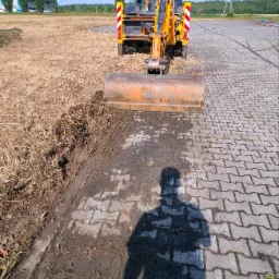 Żółta koparko-ładowarka z operatorem w kabinie, usuwająca resztki trawy i ziemi z kostki brukowej w Trzebnicy, widoczny cień osoby robiącej zdjęcie.