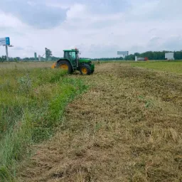 Zielony traktor John Deere z ż&oacute;łtymi kołami kosi wysoką trawę na rozległym polu w pochmurny dzień, widoczny billboard Castorama i zabudowania przemysłowe w tle.