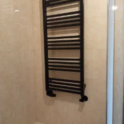 Czarny, drabinkowy grzejnik łazienkowy zamontowany na beżowych, kwadratowych płytkach. Widoczne podłączenie do instalacji.