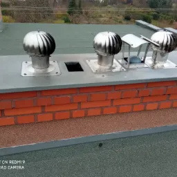 Komplet trzech metalowych nasad kominowych typu turbowent na ceglanym kominie, widok z góry na tle zielonego dachu pokrytego papą i drzew w oddali.