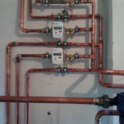 Kompleksowa instalacja z miedzianych rur z zamontowanymi licznikami ciepła, zaworami i elementami mocującymi na szarej ścianie.