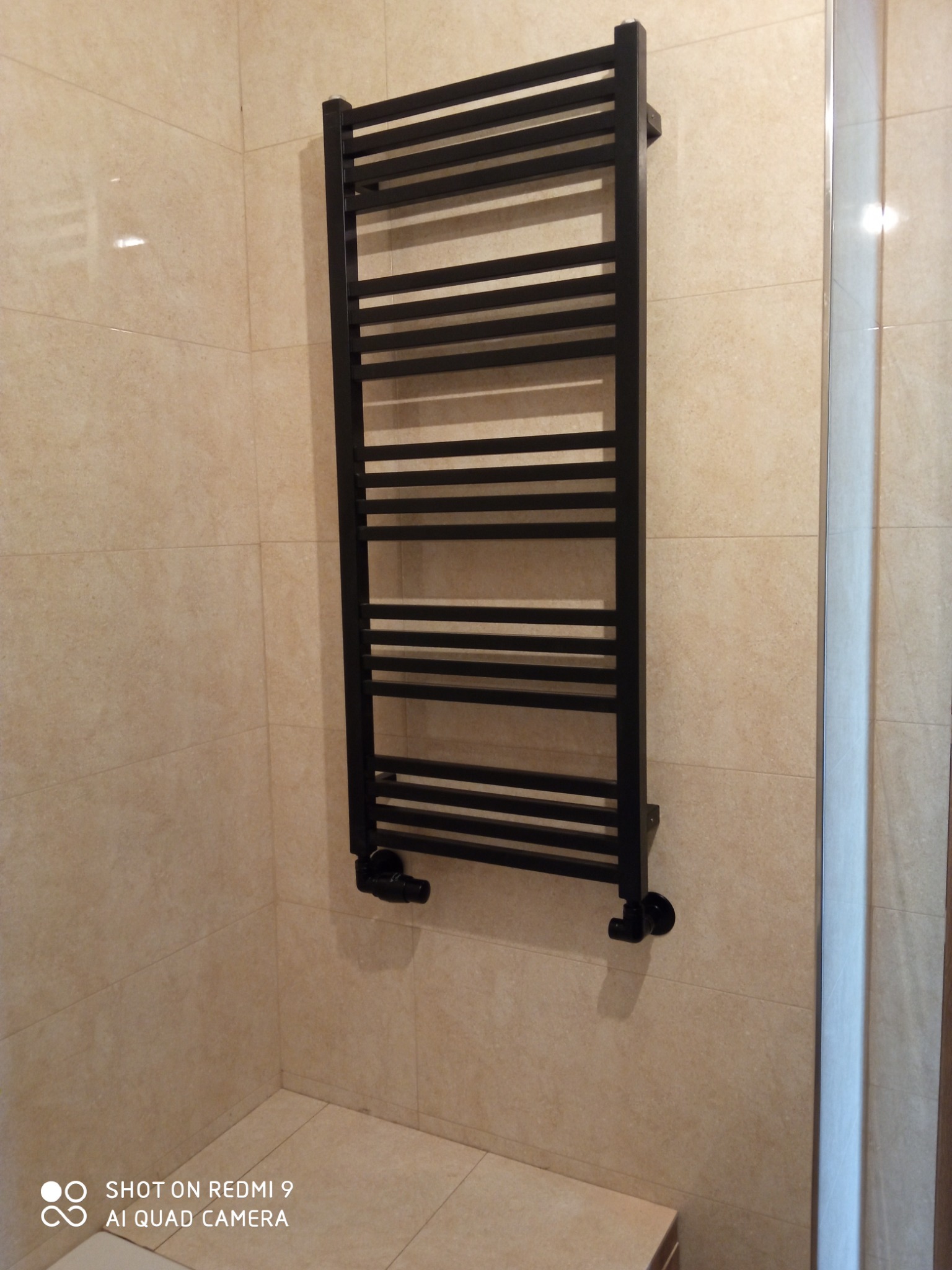 Czarny, drabinkowy grzejnik łazienkowy zamontowany na beżowych, kwadratowych płytkach. Widoczne podłączenie do instalacji.