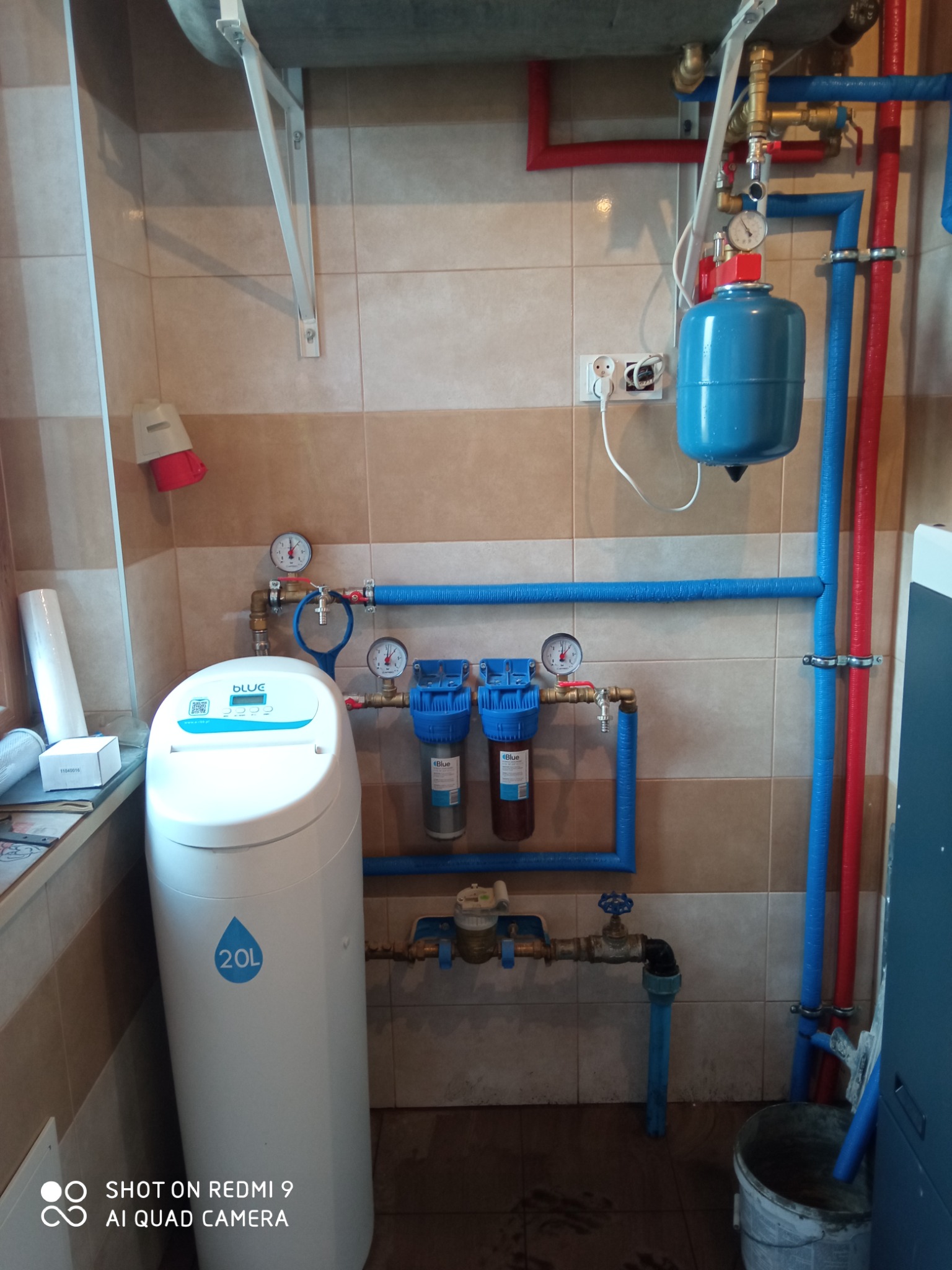 Zainstalowany system uzdatniania wody z filtrami i zbiornikiem ciśnieniowym, rury instalacyjne w kolorze niebieskim i czerwonym, manometry na ścianie wyłożonej płytkami w odcieniach beżu i brązu.