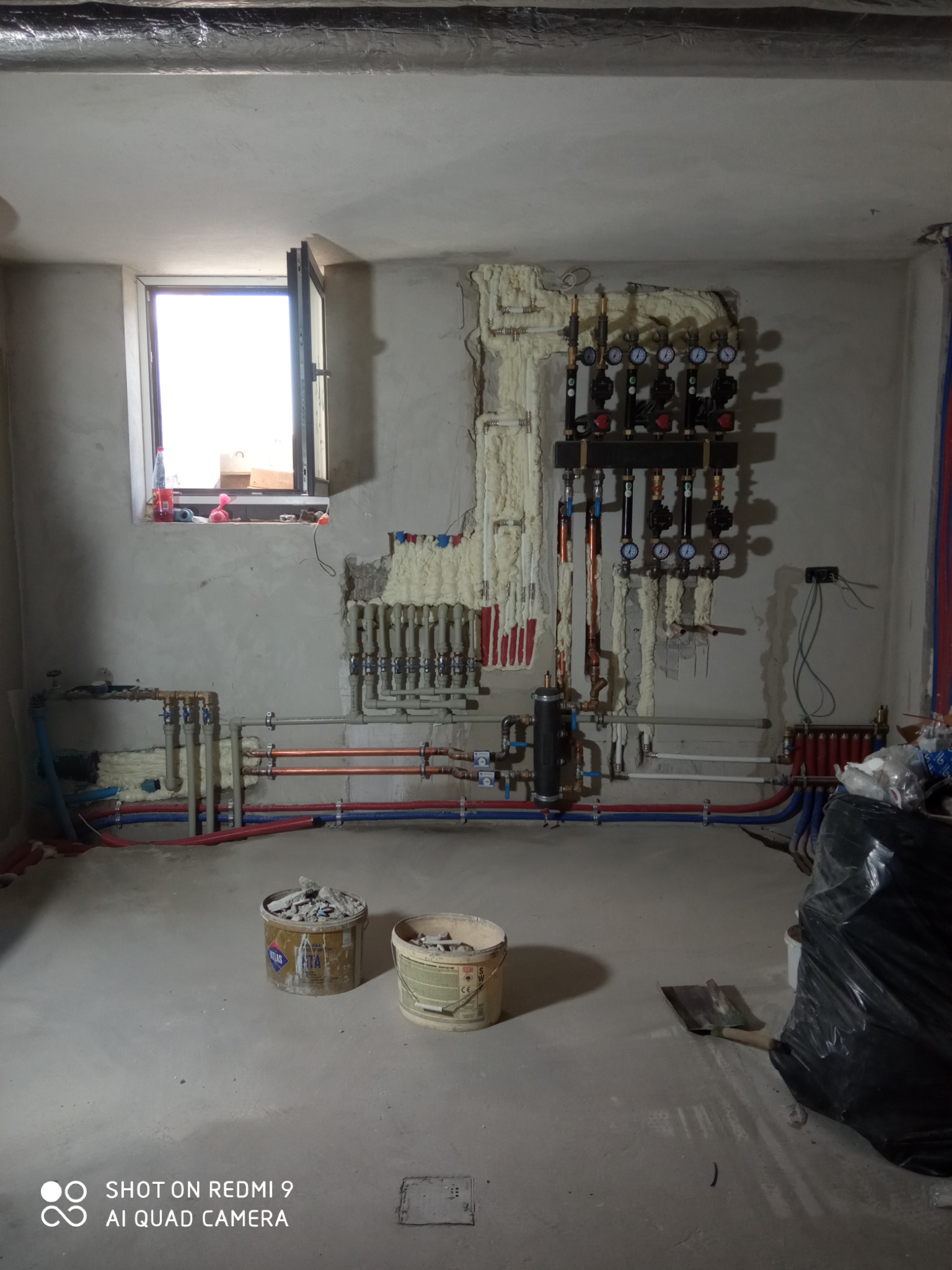 Surowy stan piwnicy w trakcie instalacji: widoczne rury grzewcze i hydrauliczne, rozdzielacz z manometrami, częściowo zaizolowane ściany, dwa wiadra z resztkami gruzu i łopata.