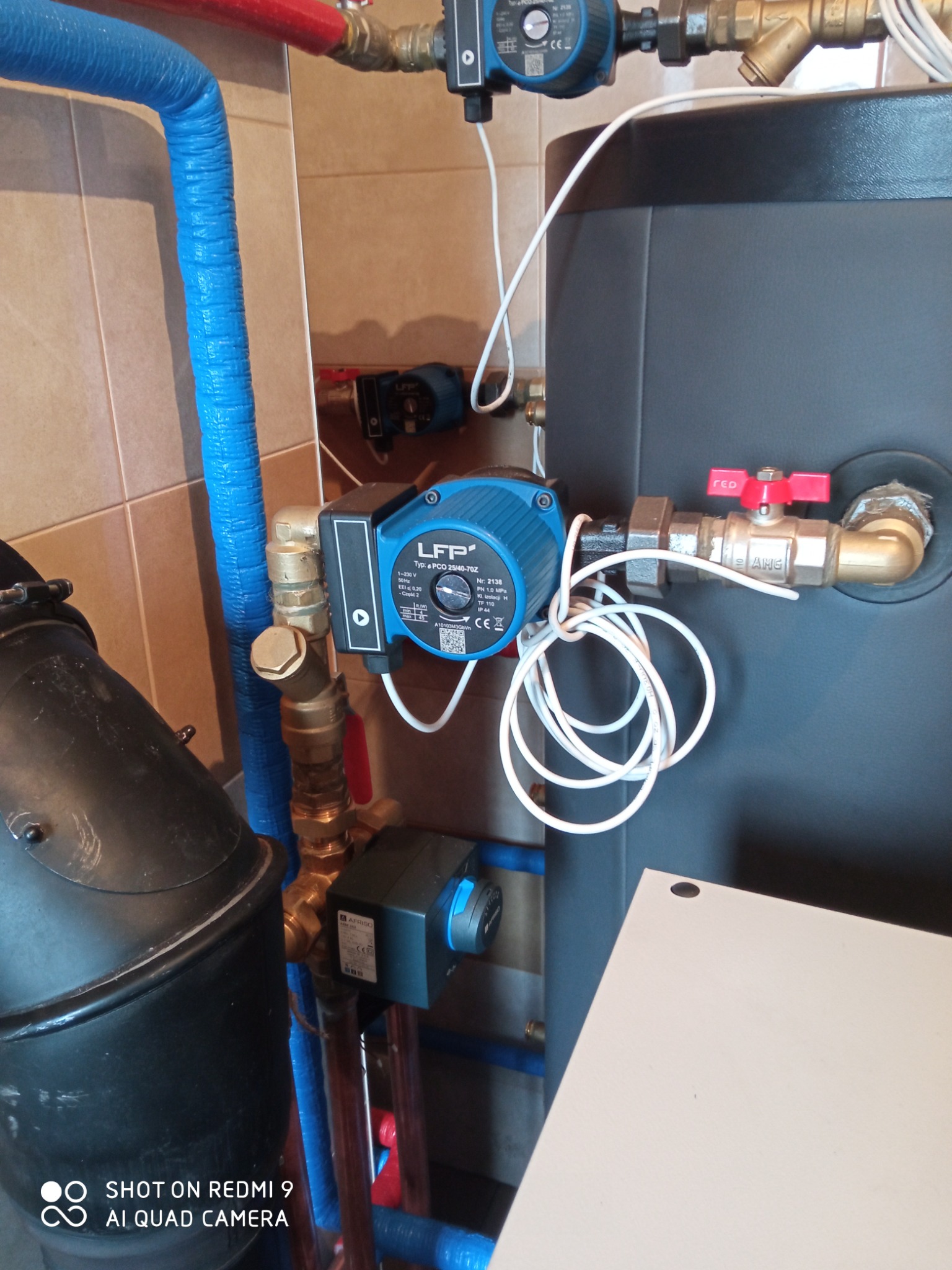 Pompa obiegowa LFP podłączona do instalacji grzewczej z izolowanymi rurami w kolorze niebieskim i czerwonym, widoczne zawory z czerwonymi pokrętłami i element kotła grzewczego w tle.