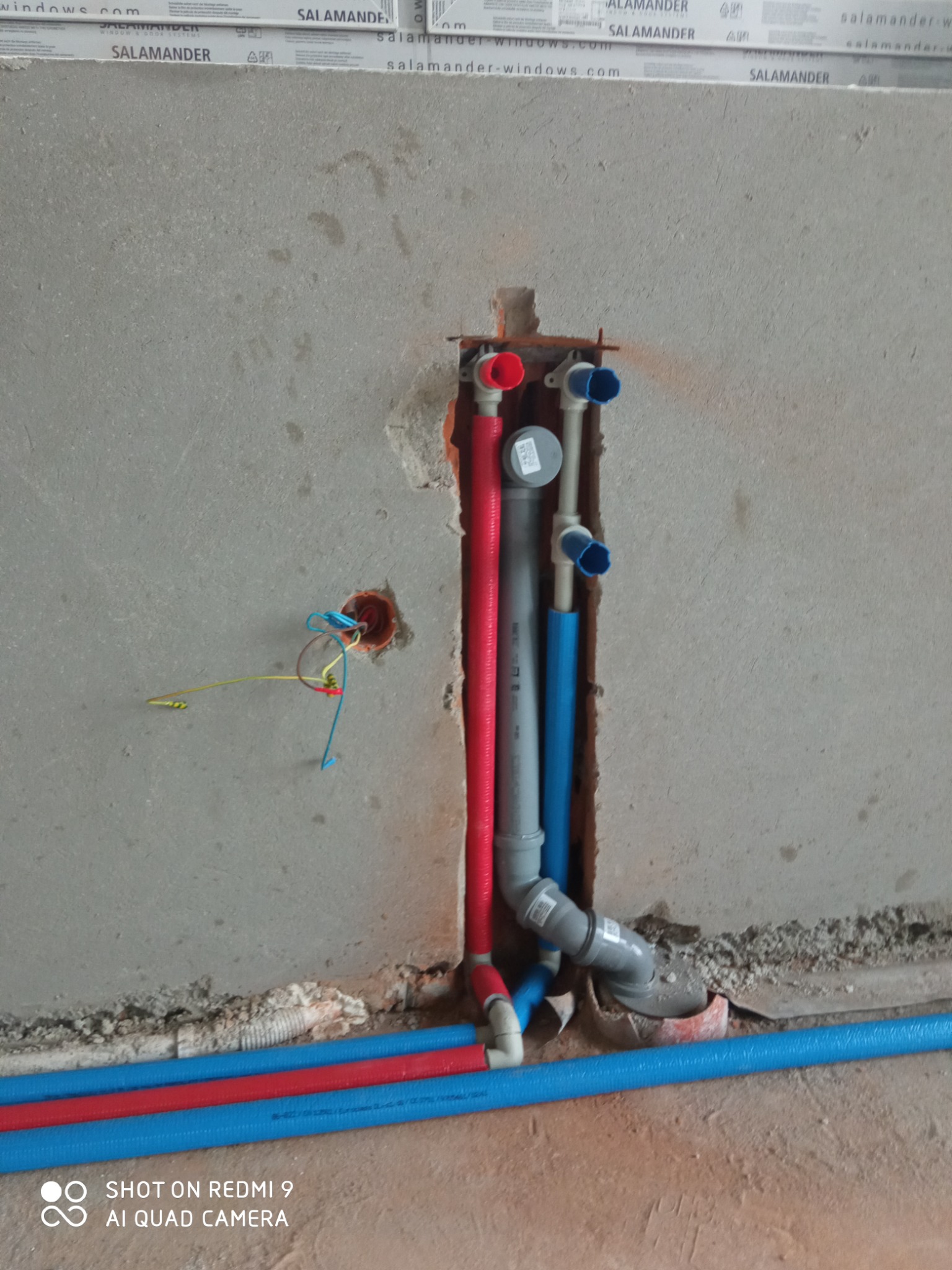 Fragment ściany w trakcie prac hydraulicznych: widoczne pionowe rury (czerwona, szara i niebieska) wystające ze ściany, z podłączeniami zakończonymi zaślepkami, oraz poziome rury na podłodze, obok...