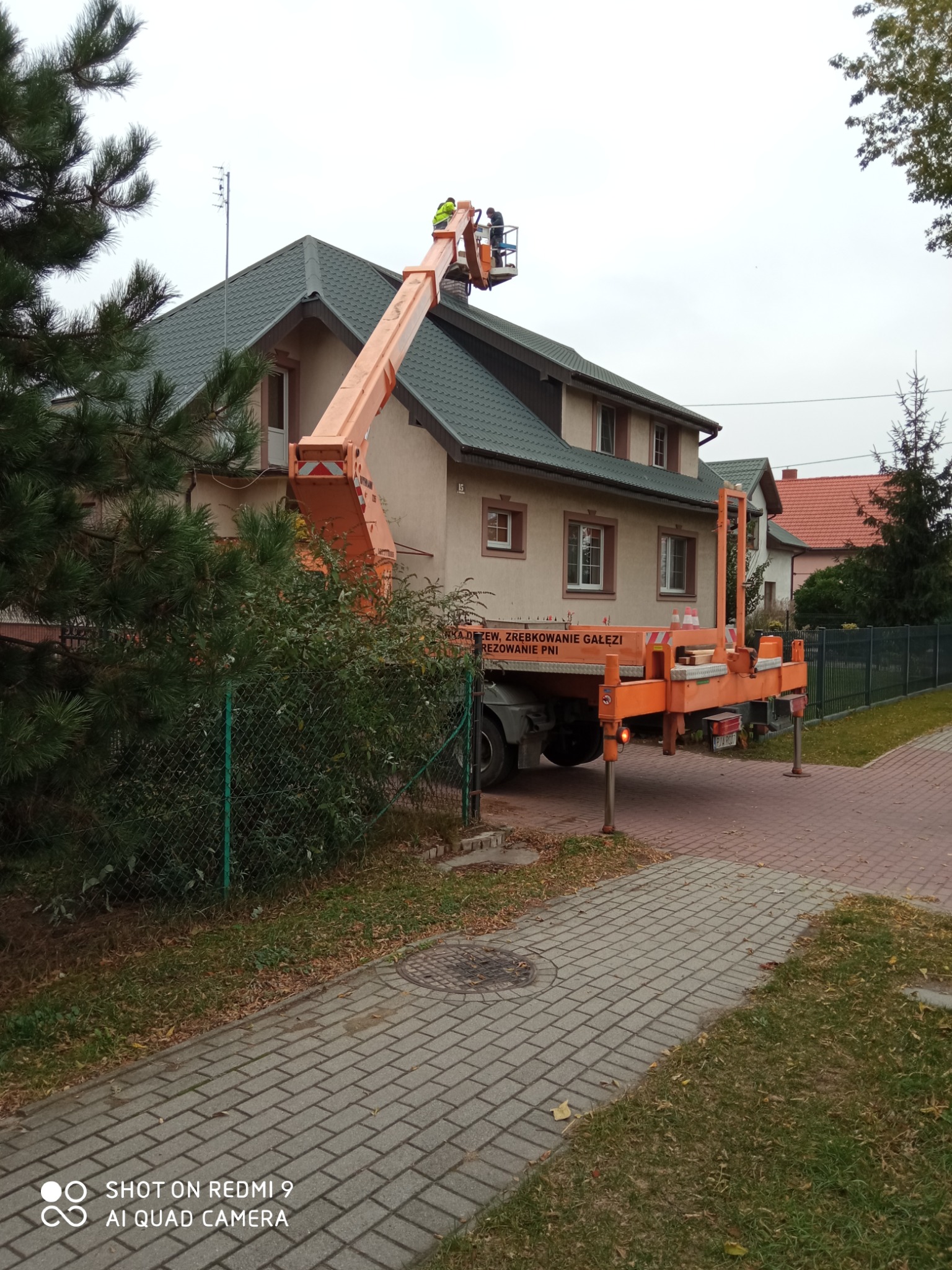 Pracownicy w pomarańczowym podnośniku koszowym na tle domu z zielonym dachem, prawdopodobnie podczas prac związanych z pielęgnacją drzew lub instalacji anteny.