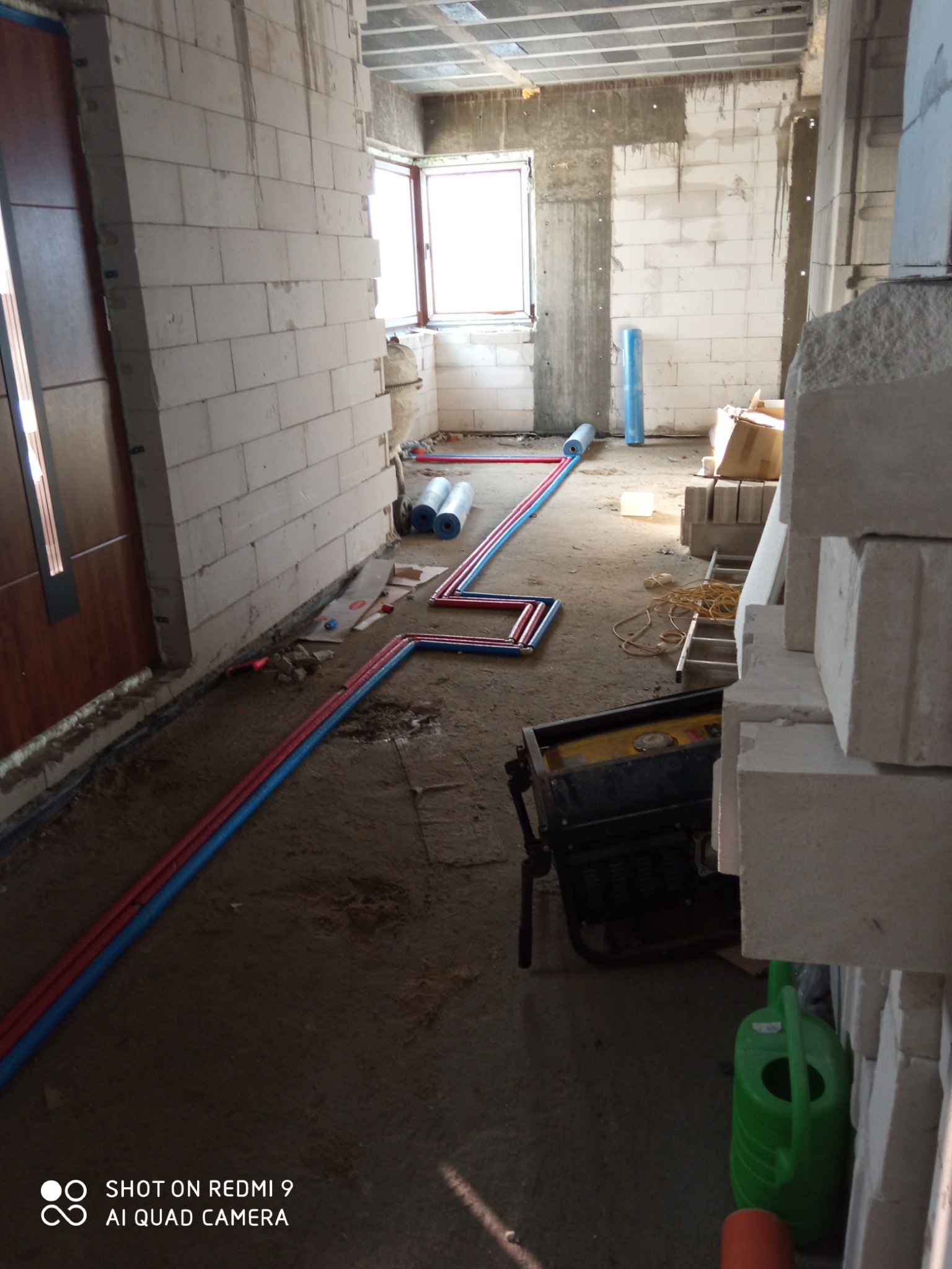 Wnętrze w trakcie prac instalacyjnych: ułożone rury w kolorach czerwonym i niebieskim na podłodze, widoczne materiały budowlane, generator prądu i zielona konewka.