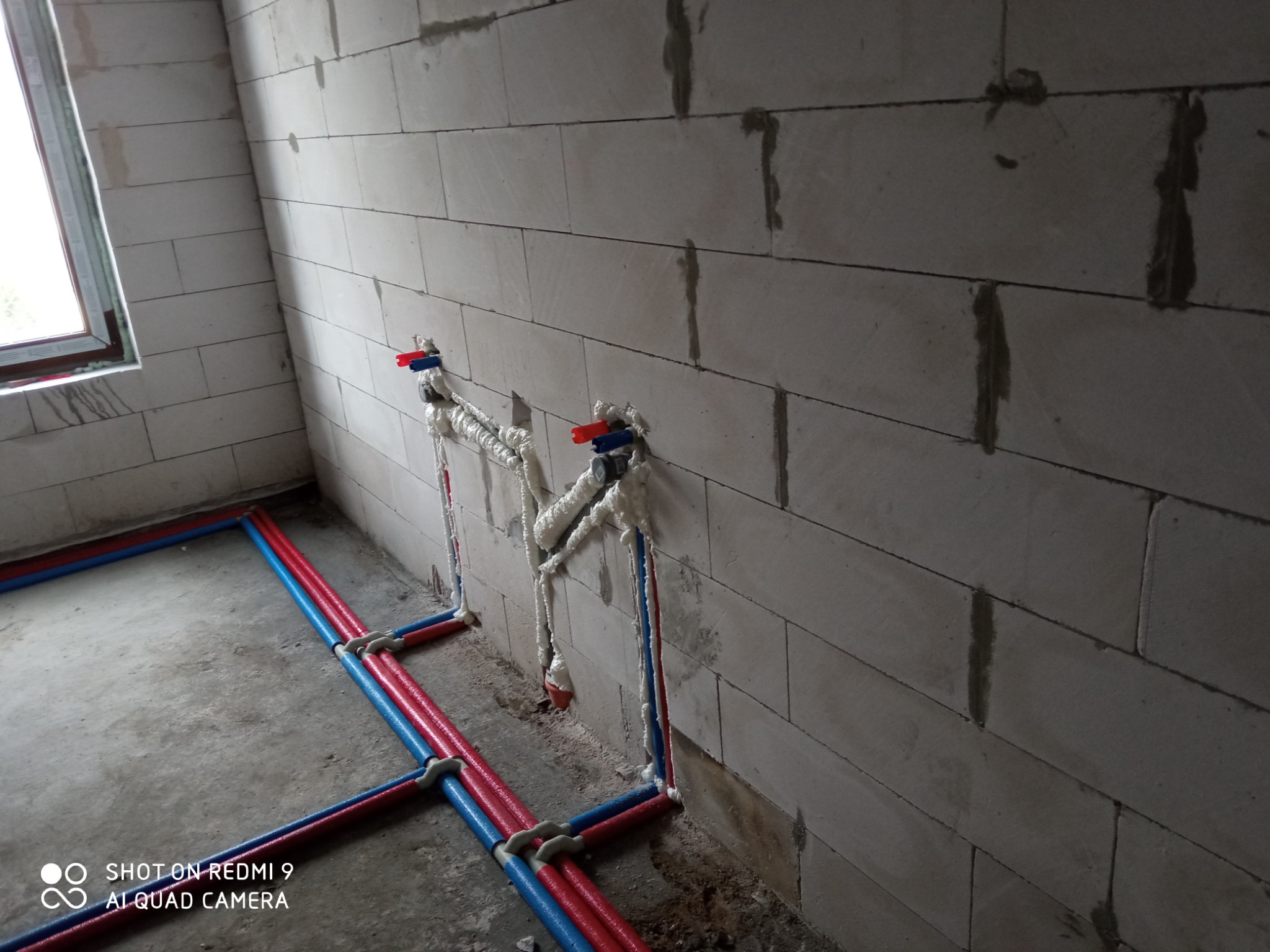 Wewnętrzne ściany z betonu komórkowego z widocznymi rurami instalacji wodnej w kolorze czerwonym i niebieskim, częściowo zabezpieczonymi pianką montażową, w surowym stanie wykończenia.