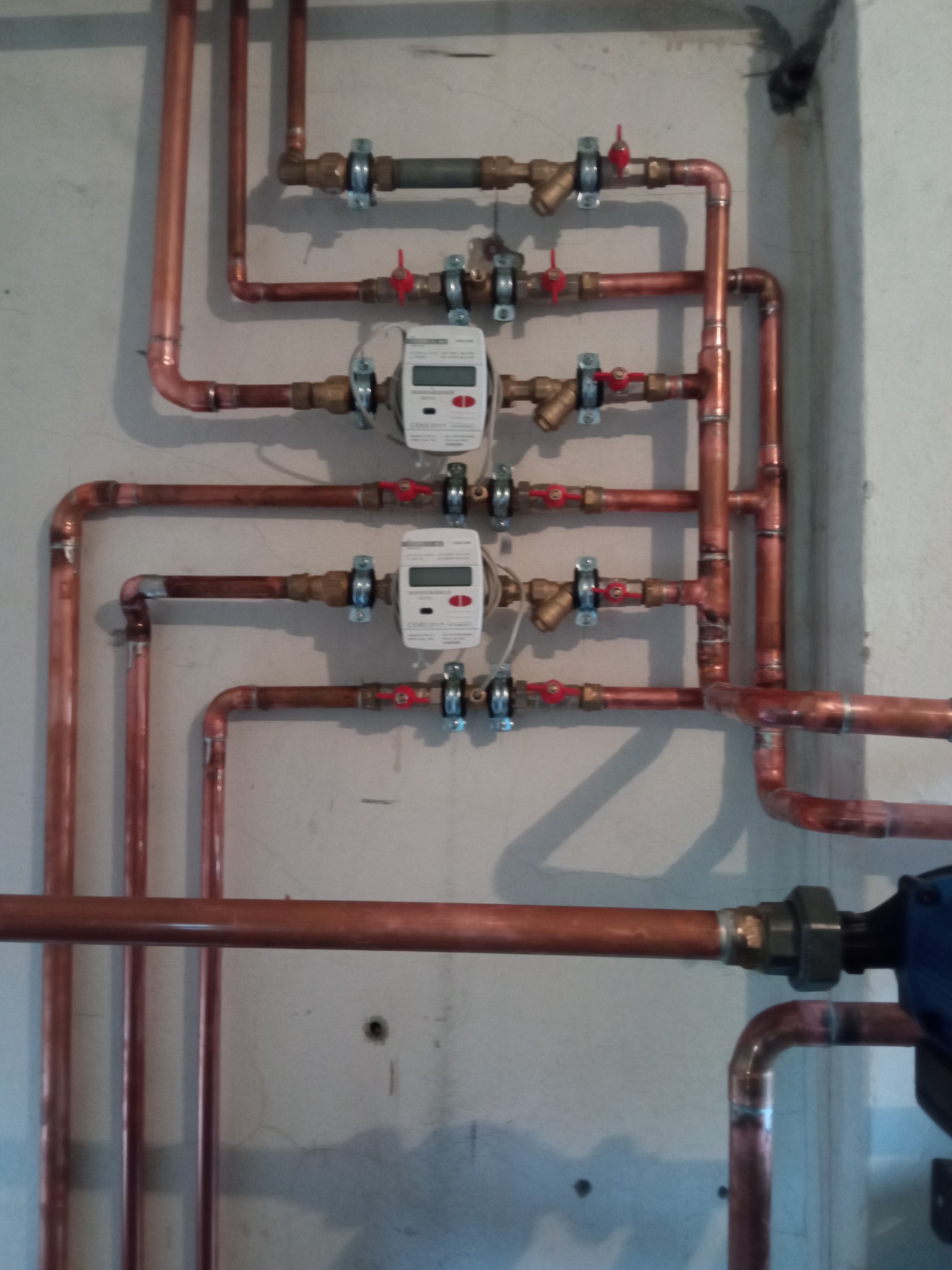 Kompleksowa instalacja z miedzianych rur z zamontowanymi licznikami ciepła, zaworami i elementami mocującymi na szarej ścianie.