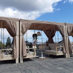 LUKAS USLUGI OGÓLNOBUDOWLANE - Zadaszone huśtawki z beżowymi zasłonami i wzorzystymi poduszkami na tarasie z szarych płyt, widok na miasto w tle.