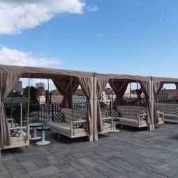 LUKAS USLUGI OGÓLNOBUDOWLANE - Zadaszone, drewniane sofy z zasłonami na tarasie z widokiem na miasto w tle.