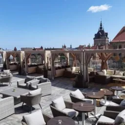 Umeblowany taras na dachu z widokiem na miasto Gdańsk, zadaszone strefy relaksu z beżowymi zasłonami, rattanowe meble wypoczynkowe, rośliny w donicach, błękitne niebo.