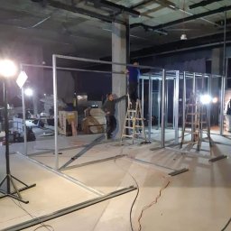 LUKAS USLUGI OGÓLNOBUDOWLANE - Montaż metalowego szkieletu ścianki działowej z płyt karton gips w surowym wnętrzu biurowym z widocznymi instalacjami pod sufitem, dwóch pracowników na drabinach, oświetlenie budowlane.