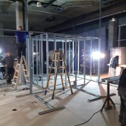 LUKAS USLUGI OGÓLNOBUDOWLANE - Konstrukcja szkieletowa z profili metalowych w trakcie montażu w surowym wnętrzu, dwóch pracowników na drabinach, widoczne instalacje pod sufitem.