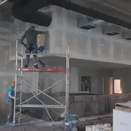 LUKAS USLUGI OGÓLNOBUDOWLANE - Praca w Gdańsku: Monter na rusztowaniu szlifuje ścianę z płyt kartonowo-gipsowych w surowym wnętrzu biurowym przed malowaniem.