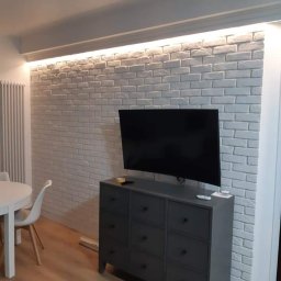 LUKAS USLUGI OGÓLNOBUDOWLANE - Jasnoszara ściana z cegły dekoracyjnej z podświetleniem LED w nowoczesnym salonie, szara komoda pod telewizorem, podłoga laminowana, białe krzesła i stół w tle.