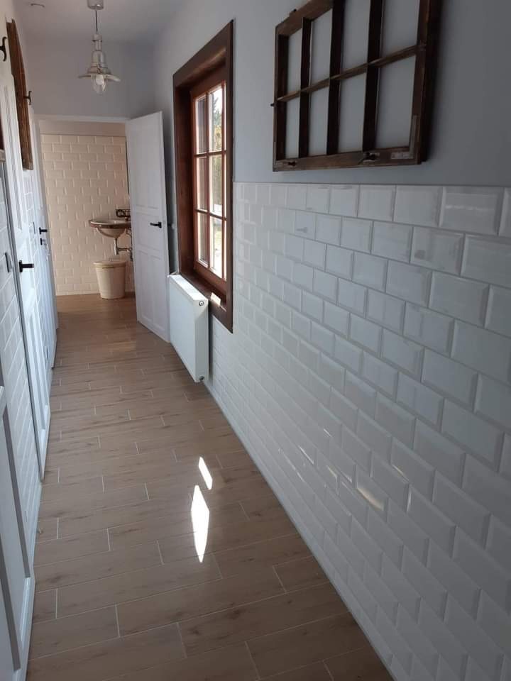 Korytarz z białymi, metrowymi płytkami na ścianie, drewnianą podłogą imitującą panele, oknem z ciemną ramą i dekoracyjną, drewnianą ramą okienną na ścianie.