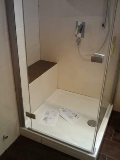 Kabina prysznicowa z siedziskiem z ciemnego drewna, biały brodzik z widocznymi instrukcjami montażu, chromowana armatura i szklane drzwi.