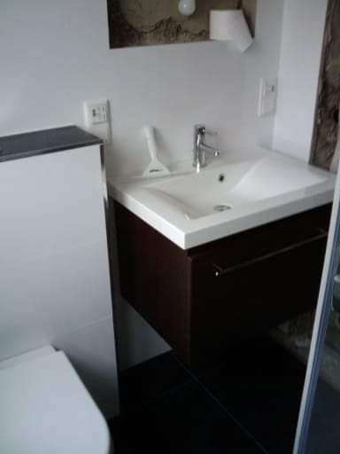 Nowoczesna umywalka z szafką w kolorze ciemnego drewna, biała ceramika, czarne płytki podłogowe, akcesoria łazienkowe i fragment toalety widoczne w kadrze.