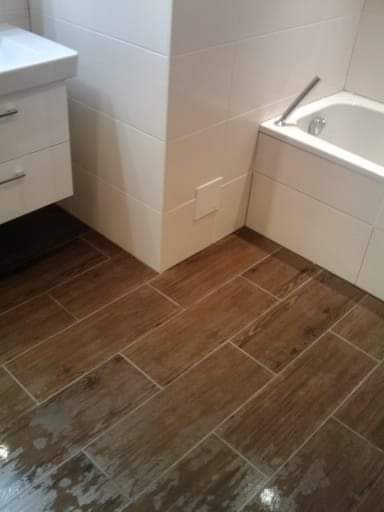 Ukończona łazienka z brązowymi płytkami imitującymi drewno na podłodze, białe płytki na ścianach, biała umywalka z szafką i fragment wanny.