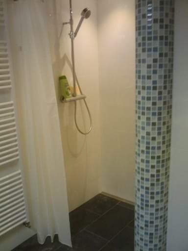 Nowoczesna kabina prysznicowa z kolumną wyłożoną mozaiką w odcieniach szarości i błękitu, widoczny biały grzejnik drabinkowy i czarna podłoga z płytek.