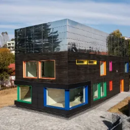 Nowoczesny dom jednorodzinny z drewnianą elewacją i górną kondygnacją wykończoną lustrzanymi panelami, okna w kolorowych ramach (zielona, niebieska, pomarańczowa, różowa, żółta), otoczenie z kostki...