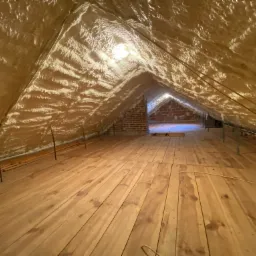 Poddasze ocieplone pianką, widok od podłogi w kierunku komina, widoczne drewniane deski podłogowe i nierówna powierzchnia pianki na skosach dachu.