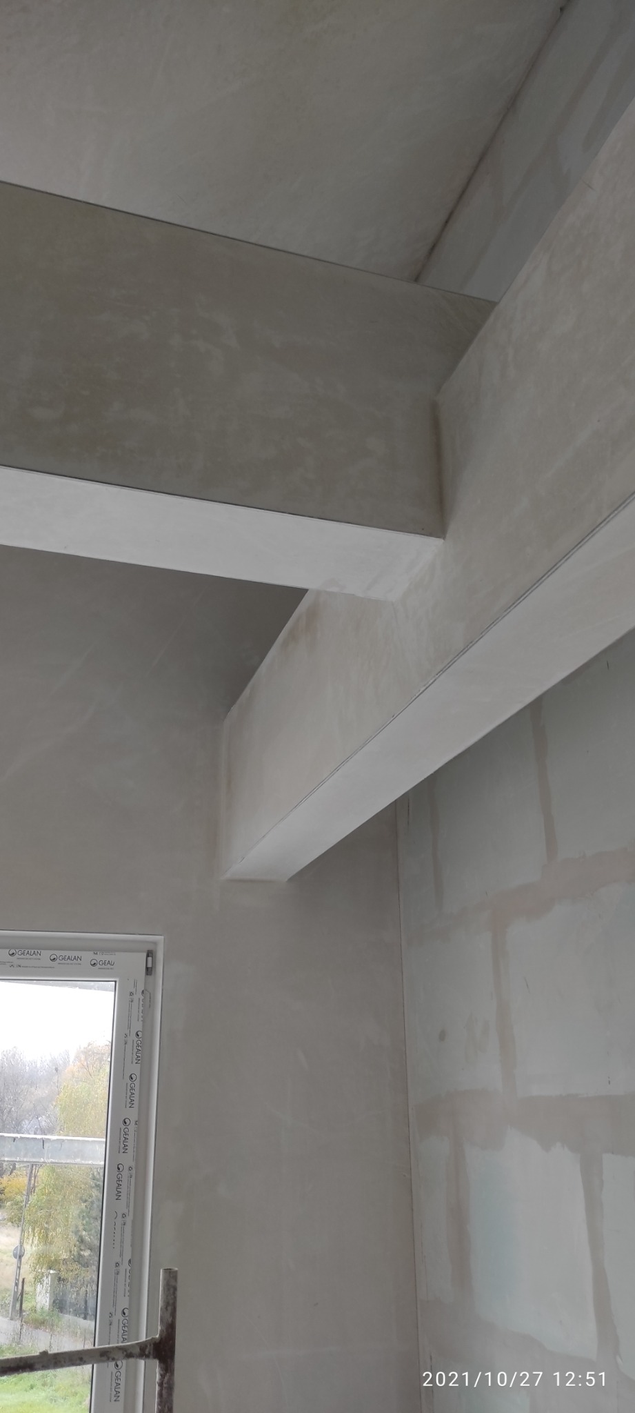 Świeżo położone tynki maszynowe na ścianach i suficie, widoczne elementy konstrukcyjne budynku oraz rama okienna z widokiem na zewnątrz.